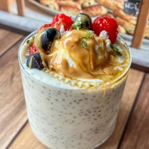 A glass of chia pudding topped with berries, blueberries, peanut butter, honey, and fresh herbs, set on a wooden table with pastries visible in the background.