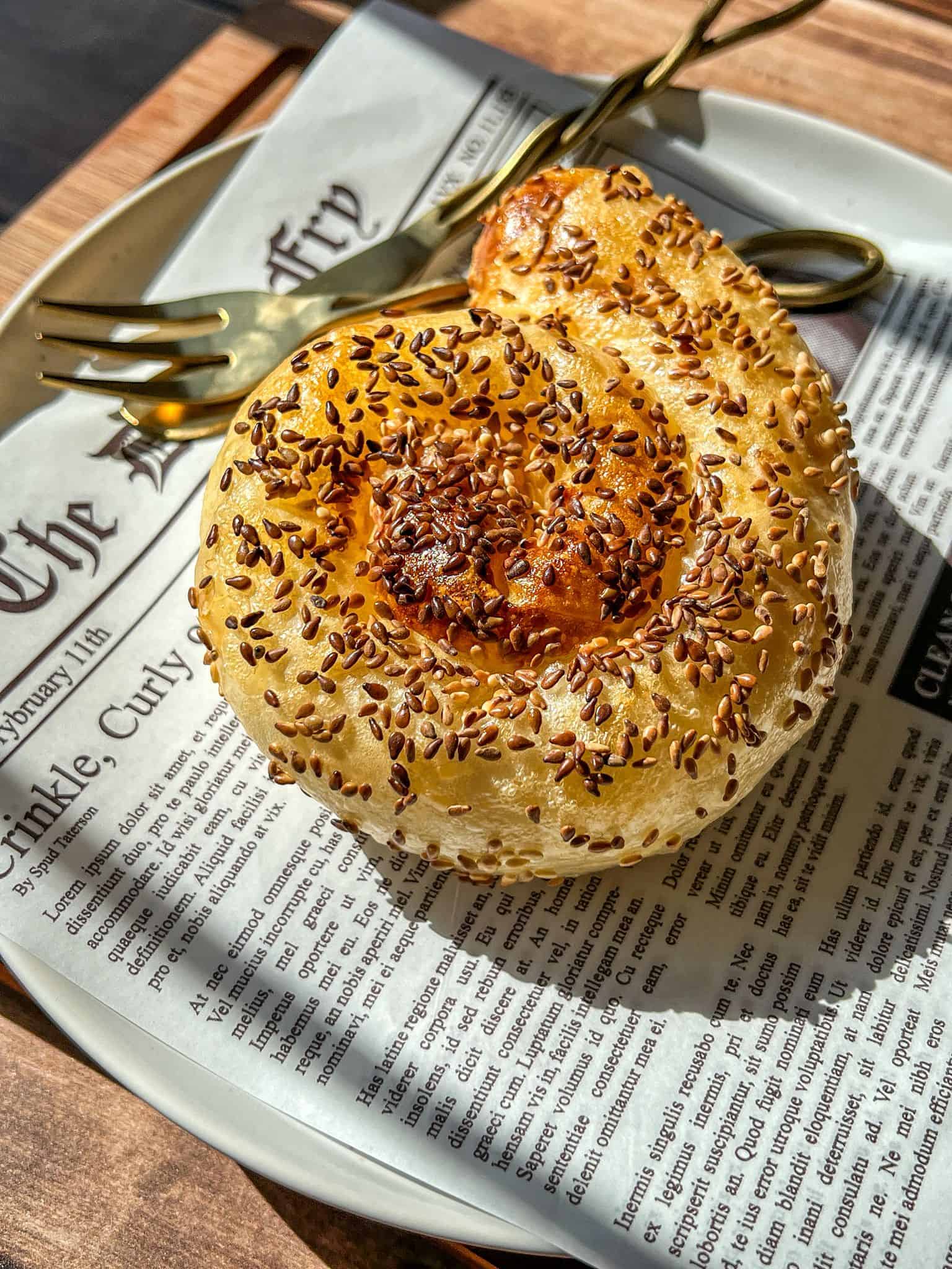 A golden, spiral-shaped pastry topped with flax seeds sits on a white plate lined with newspaper print, next to a gold fork and knife, in bright natural sunlight.