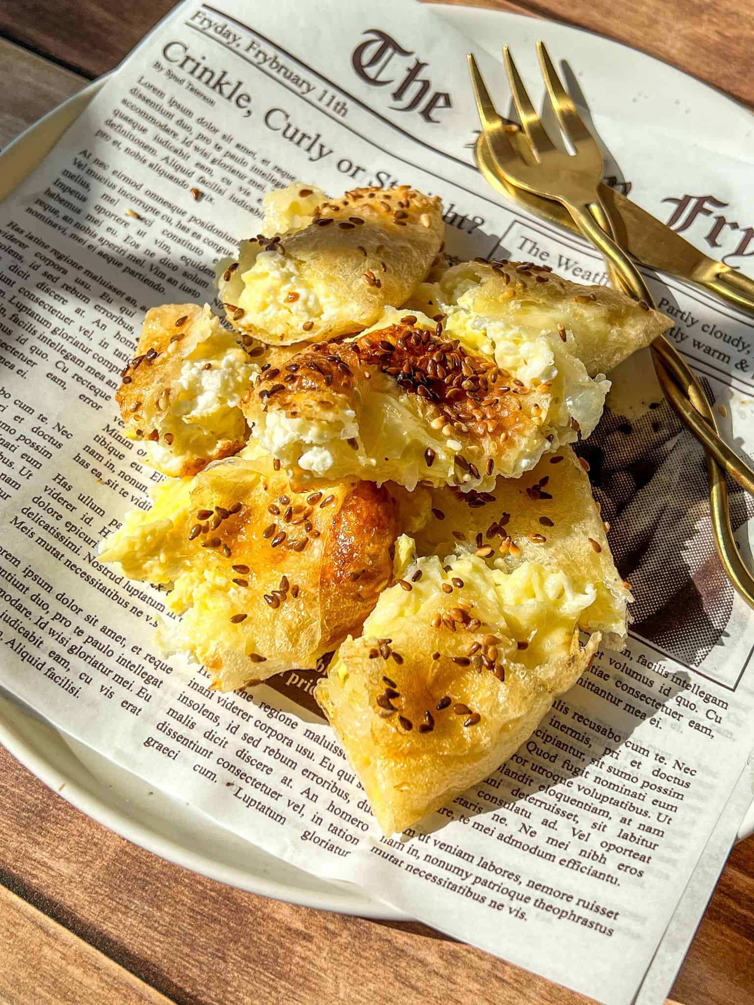 Golden, flaky bread topped with sesame seeds and cheese sits on a newspaper-lined plate with two gold forks, illuminated by warm sunlight.