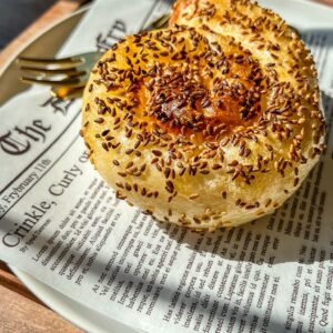A round, golden pastry topped with sesame seeds sits on a white plate lined with newspaper-print parchment. A gold fork rests beside it, and sunlight casts soft shadows across the scene.