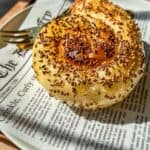 A golden, spiral-shaped pastry topped with sesame seeds sits on a plate lined with newspaper-style paper, with a fork beside it. Sunlight highlights the flaky texture of the pastry.
