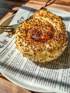 A golden, spiral-shaped pastry topped with sesame seeds sits on a plate lined with newspaper-style paper, with a fork beside it. Sunlight highlights the flaky texture of the pastry.