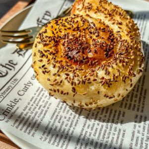 A golden, spiral-shaped pastry topped with sesame seeds sits on a plate lined with newspaper-style paper, with a fork beside it. Sunlight highlights the flaky texture of the pastry.