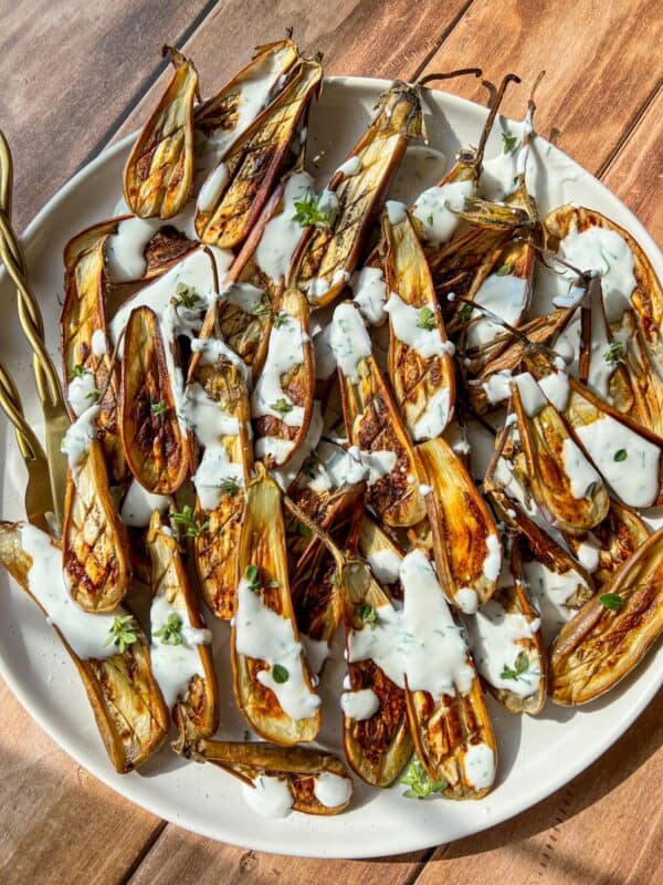 A large white plate of roasted baby eggplants, halved and topped with a drizzle of white yogurt sauce and chopped herbs, sits on a wooden table in sunlight. Two gold-toned serving forks rest on the plate's edge.