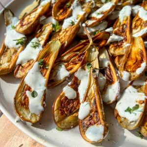 A plate of roasted baby eggplant halves topped with a drizzle of creamy white sauce and garnished with fresh green herbs, set on a wooden surface in natural sunlight.