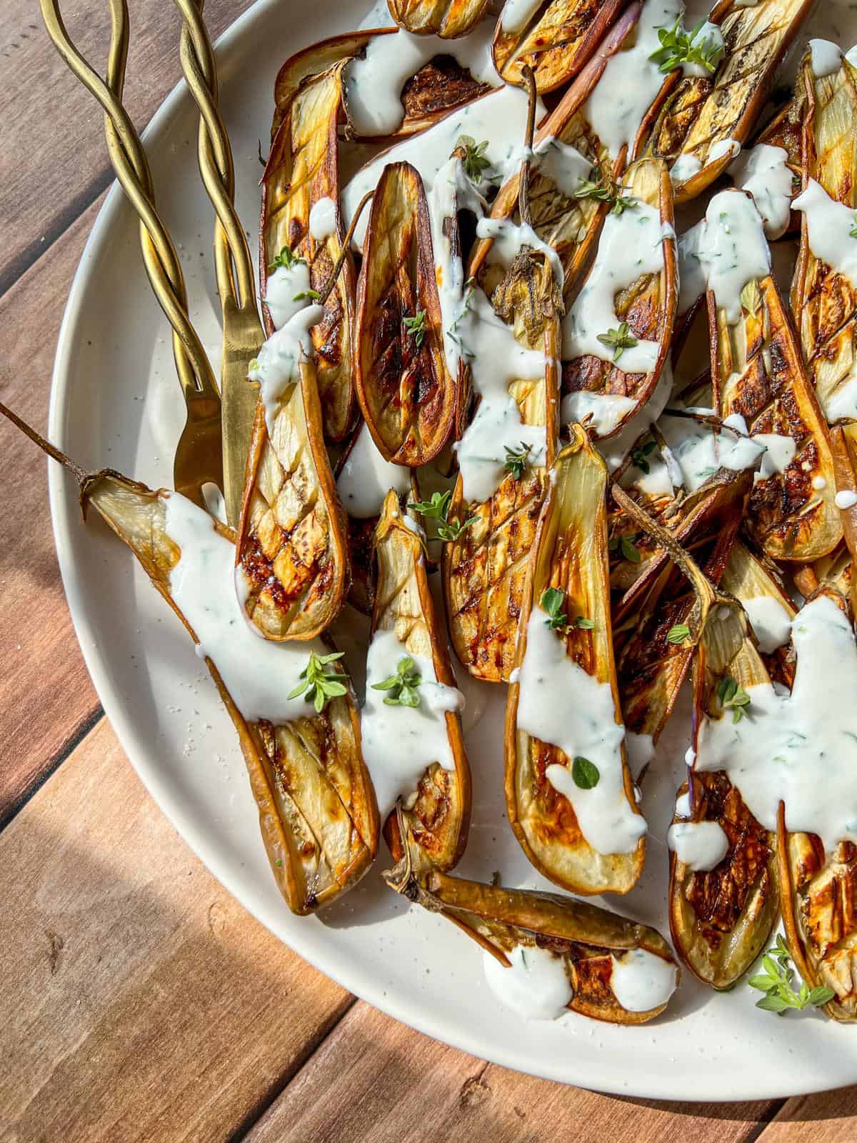 Grilled eggplant halves arranged on a white plate, drizzled with creamy white sauce and garnished with fresh herbs, with gold forks placed on the side.