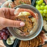 A hand holds a cracker sandwich with cheese and prosciutto above a tray with crackers, grapes, and cheese on a wooden table.