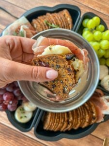 A hand holds a cracker sandwich with cheese and prosciutto above a tray with crackers, grapes, and cheese on a wooden table.