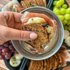 A hand holds a cracker sandwich with cheese and prosciutto above a tray with crackers, grapes, and cheese on a wooden table.