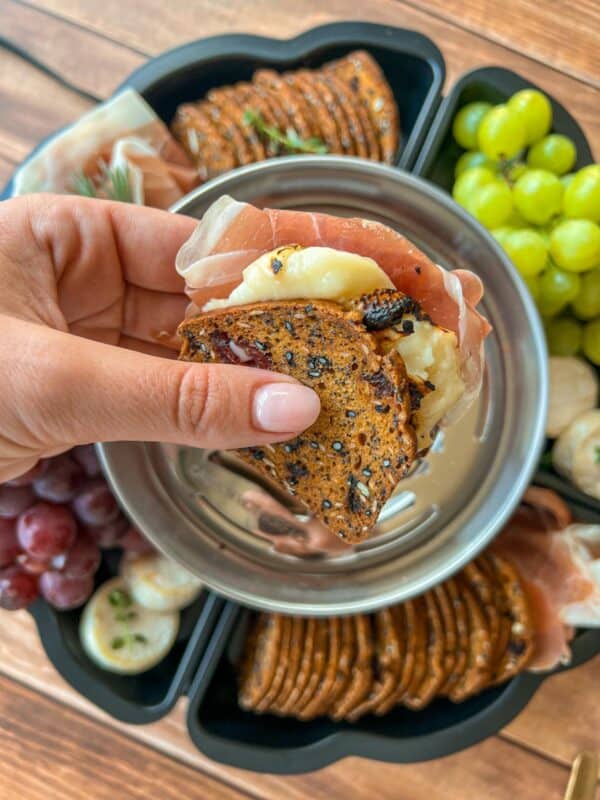 A hand holds a cracker sandwich with cheese and prosciutto above a tray with crackers, grapes, and cheese on a wooden table.