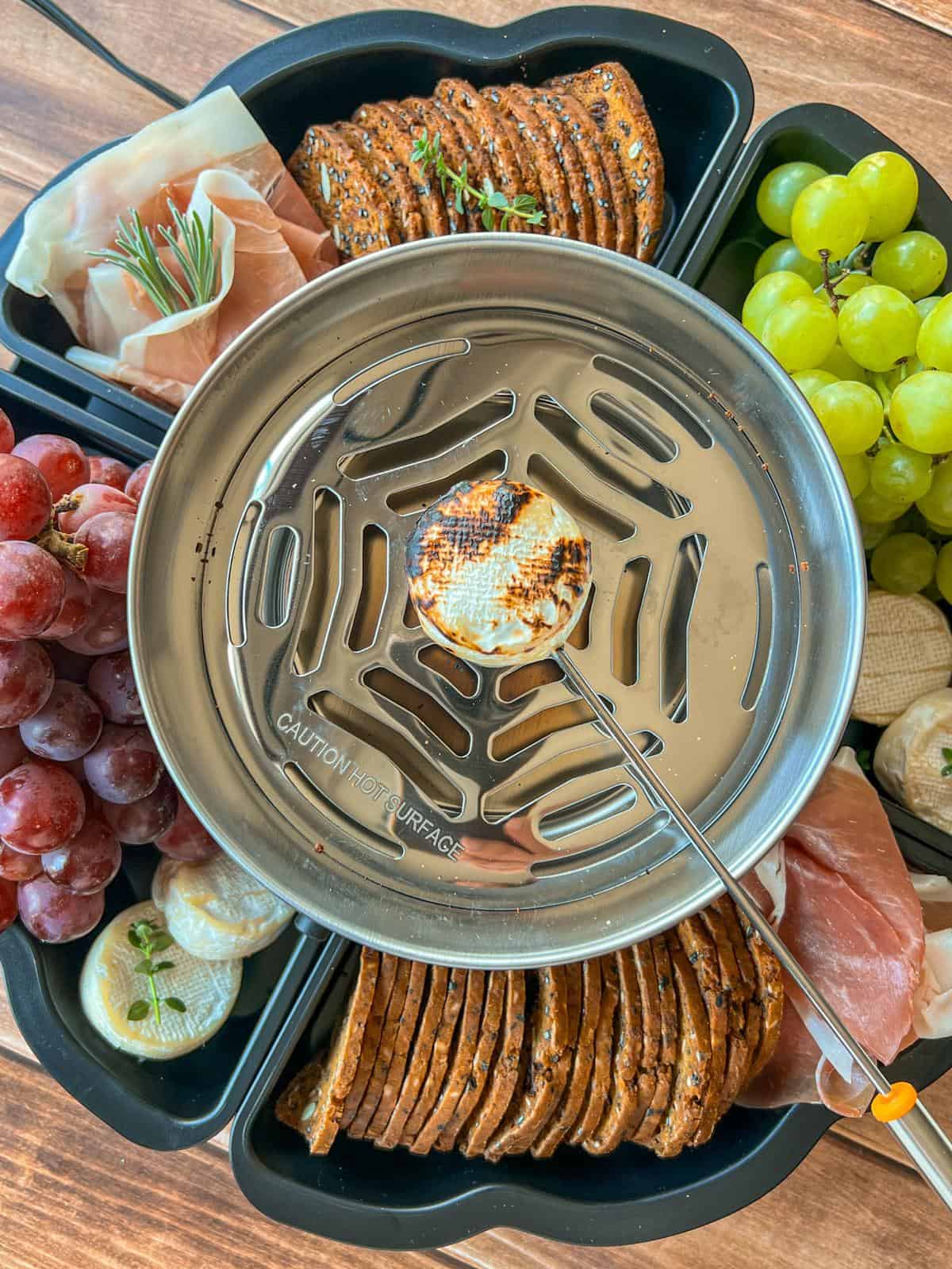 A metal burner toasts a marshmallow for Savory Smores on a skewer, surrounded by crackers, grapes, cheese, prosciutto, and herbs, all arranged in a black snack tray on a wooden surface.