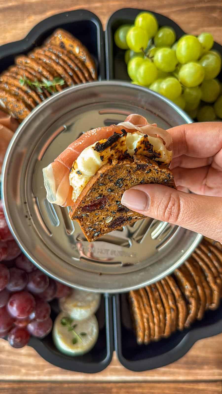 A hand holds a stack of crackers with cheese and prosciutto above a metal dish. The background shows green grapes, red grapes, crackers, and cheese displayed on a wooden surface.