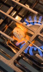 A marshmallow on a metal skewer is being roasted over a blue gas stove flame, with part of the marshmallow turning brown.