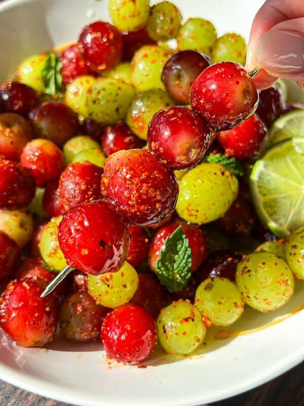 A hand holds a skewer with red and green grapes coated in chili powder above a bowl filled with more seasoned grapes, lime slices, and mint leaves on a wooden surface.