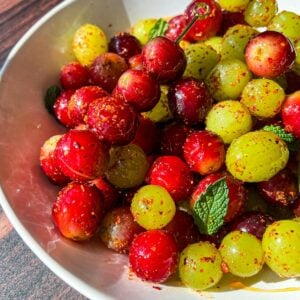 A white bowl filled with red and green grapes coated in chili powder and seasoning, garnished with fresh mint leaves, placed on a wooden surface.