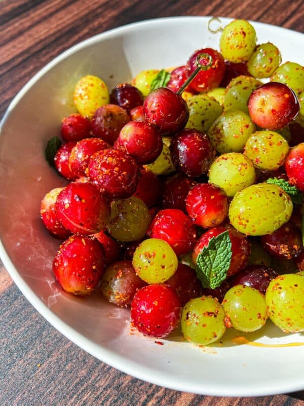 A white bowl filled with red and green grapes coated in chili powder and seasoning, garnished with fresh mint leaves, placed on a wooden surface.