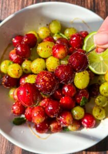 A close-up of a bowl filled with glossy red and green gooseberries coated in spices, with a lime slice visible. A hand holds a skewer with three spiced gooseberries above the bowl.
