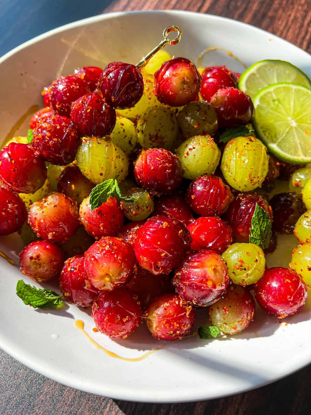A white bowl filled with red and green gooseberries sprinkled with spices, garnished with fresh mint leaves and lime slices, sitting on a dark wooden surface.
