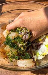 A hand mixes raw shrimp with chopped herbs, lemon zest, and seasonings in a glass bowl on a wooden surface.