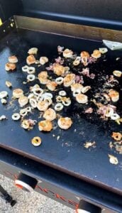 Seafood including shrimp and calamari rings cooking on a flat-top Blackstone grill with scattered seasoning and oil visible on the hot surface.