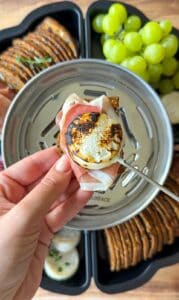 A hand holds a cracker topped with prosciutto and torched goat cheese over a serving tray with grapes, crackers, and cheese.