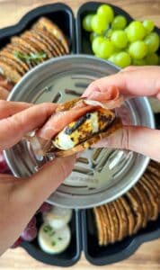 Close-up of hands assembling a snack with grilled cheese, prosciutto, and crackers, with a tray of grapes, crackers, and other snacks in the background.