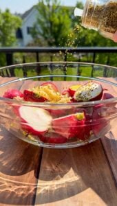 A glass bowl filled with halved radishes, lemon zest, and olive oil sits on a wooden table outdoors, while dried herbs are being sprinkled from a jar above the bowl. Greenery and a house appear in the blurred background.