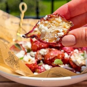 A hand holds a beet chip topped with crumbled cheese above a bowl filled with more beet chips and cheese, with a blurred green outdoor background.