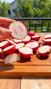 A hand holds a sliced radish with a pink and white center above a wooden board covered with more halved radishes. The background shows a sunny outdoor scene with greenery and a house.