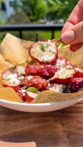 A hand holds a roasted cherry tomato topped with crumbled cheese and herbs above a bowl filled with similar tomatoes, all resting on printed parchment paper, with greenery visible in the background.