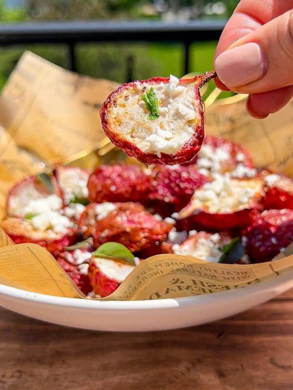 A hand holds a roasted cherry tomato topped with crumbled cheese and herbs above a bowl filled with similar tomatoes, all resting on printed parchment paper, with greenery visible in the background.