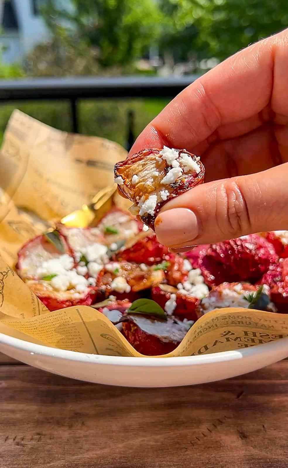 A hand holding a sliced roasted air fryer radish topped with crumbled cheese over a bowl of roasted beets garnished with cheese and herbs, served on paper-lined dish outdoors.