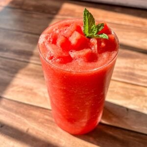 A glass of bright red watermelon smoothie topped with small watermelon cubes and a sprig of fresh mint, placed on a wooden table in natural sunlight.