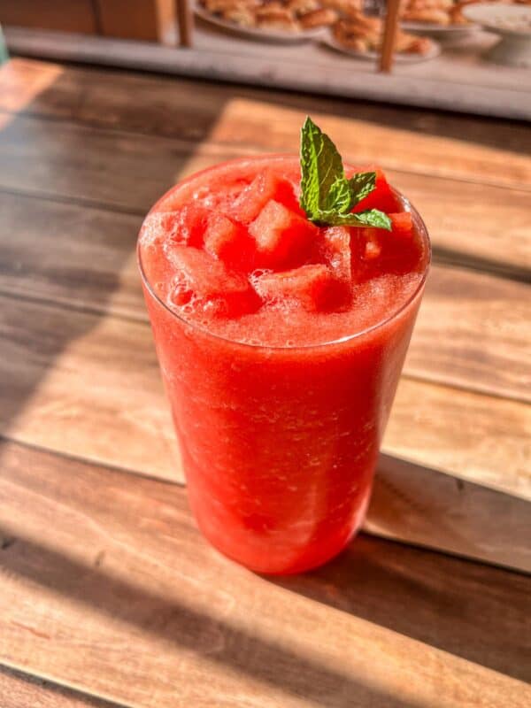 A glass of bright red watermelon smoothie topped with small watermelon cubes and a sprig of fresh mint, placed on a wooden table in natural sunlight.