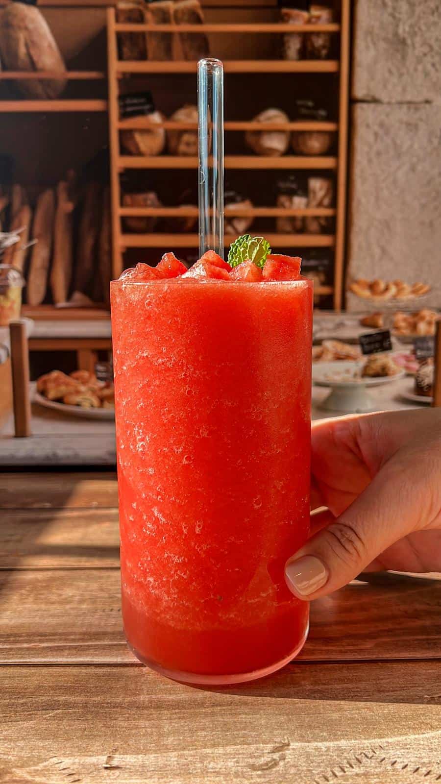 A hand holds a tall glass of bright red, frozen watermelon drink garnished with a mint leaf. Shelves with bread and pastries are blurred in the background on a wooden table.