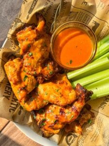 A plate of saucy grilled chicken wings with fresh celery sticks and a bowl of orange dipping sauce, served on printed parchment paper.