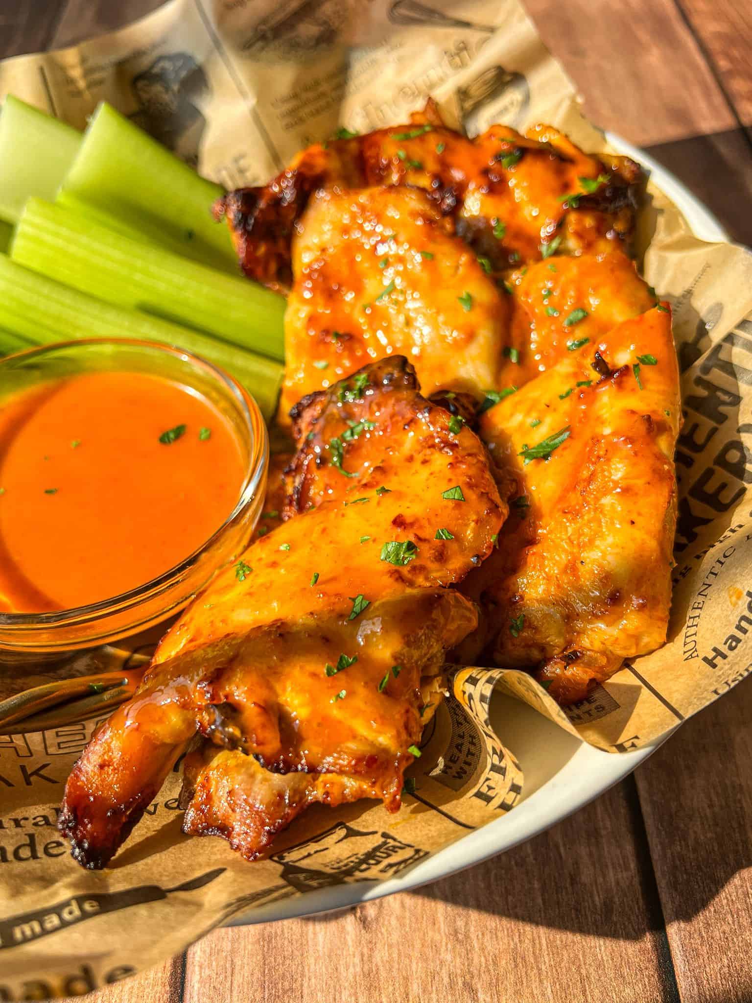 A plate of grilled buffalo chicken wings garnished with herbs, served with fresh celery sticks and a small bowl of orange buffalo sauce on brown parchment paper.