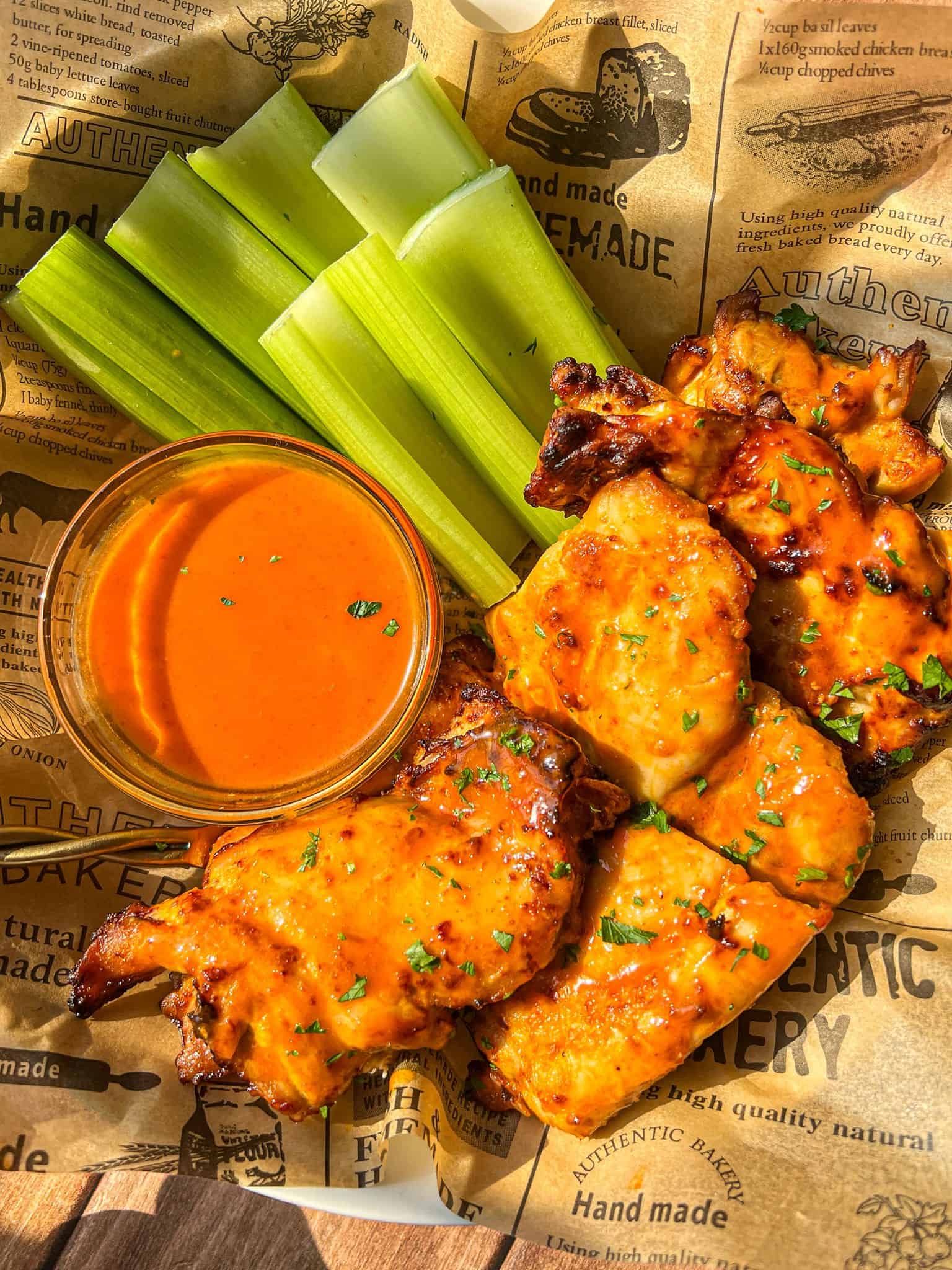 Buffalo chicken wings arranged with celery sticks and a cup of orange buffalo sauce, served on parchment-style paper with text and graphics.