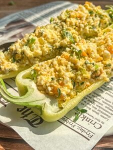 Two halved green bell peppers filled with a creamy egg salad mixture, garnished with fresh herbs, are placed on a sheet of newspaper. Sunlight highlights the vibrant colors and textures of the dish.