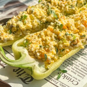 Two halved green bell peppers filled with a creamy egg salad mixture, garnished with fresh herbs, are placed on a sheet of newspaper. Sunlight highlights the vibrant colors and textures of the dish.