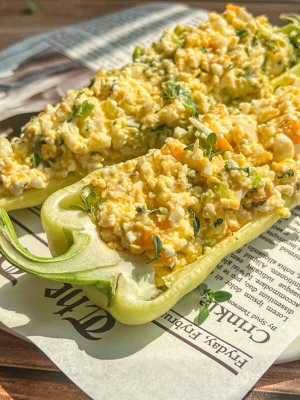 Two halved green bell peppers filled with a creamy egg salad mixture, garnished with fresh herbs, are placed on a sheet of newspaper. Sunlight highlights the vibrant colors and textures of the dish.