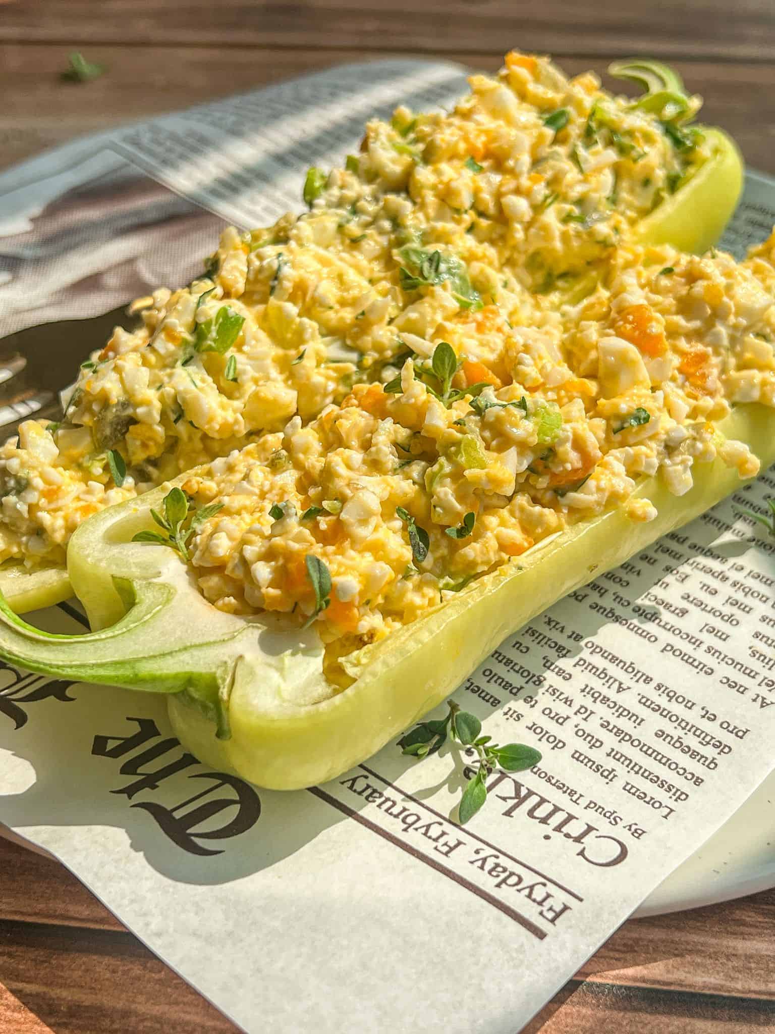 Two halved green bell peppers filled with a creamy egg salad mixture, garnished with fresh herbs, are served on a plate lined with newspaper-style parchment paper. Sunlight highlights the fresh, colorful dish.