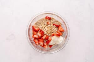 A glass bowl filled with rolled oats, chopped fresh strawberries, and a dollop of yogurt, placed on a light marble surface.
