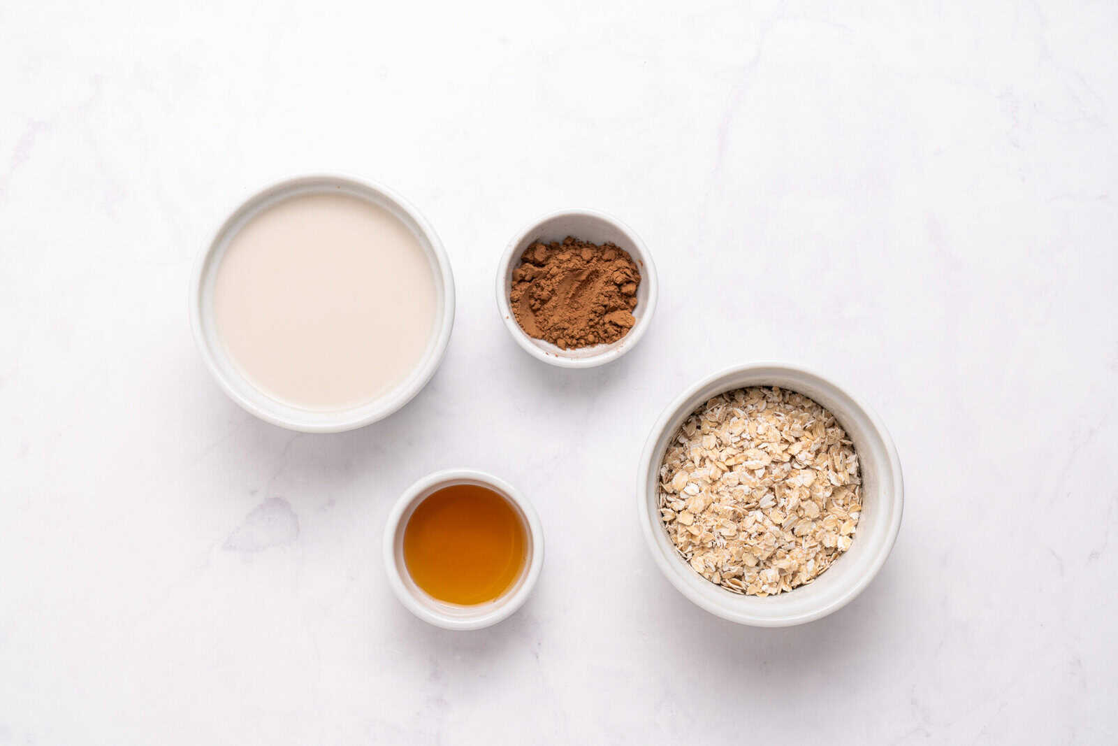 Four small bowls on a white surface containing oat milk, cocoa powder, rolled oats, and maple syrup, arranged in a flat lay style.