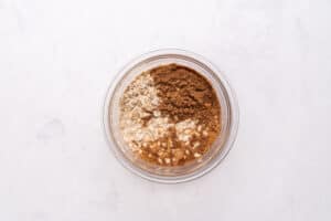 A glass bowl containing oats, cocoa powder, and other dry baking ingredients on a white marble surface, viewed from above.