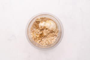 A glass bowl containing dry oats, water, and a dollop of yogurt, viewed from above on a white surface.
