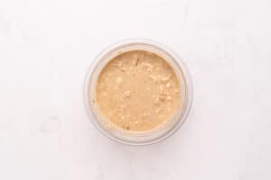 A clear bowl filled with light brown, creamy overnight oats sits on a white marble surface.