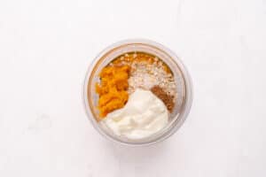 A clear glass bowl containing oats, pumpkin puree, yogurt, and spices, all neatly separated, sits on a white marble surface.