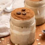Two glass jars filled with creamy tiramisu overnight oats, topped with a layer of cocoa powder and coffee beans, are placed on a wooden surface with scattered oat flakes and a spoon nearby.