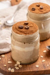 Two glass jars filled with creamy tiramisu overnight oats, topped with a layer of cocoa powder and coffee beans, are placed on a wooden surface with scattered oat flakes and a spoon nearby.